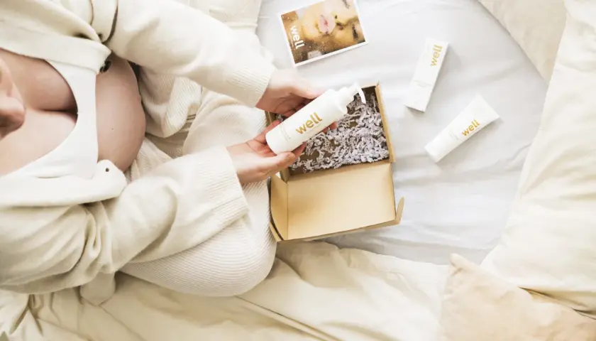 Gravid kvinna öppnar ett paket med hudvårdsprodukter. En flaska med hyaluronsyra och en kräm för bristningar är synliga.