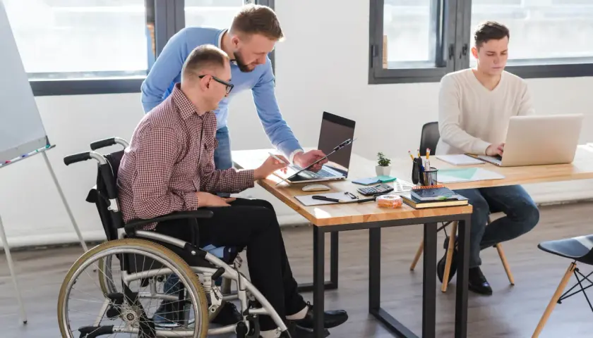 Dwóch mężczyzn pracuje przy biurku. Jeden siedzi na wózku inwalidzkim, drugi stoi obok. Trzeci mężczyzna pracuje przy laptopie przy innym biurku. To przykład efektywnego czasu pracy osoby niepełnosprawnej.