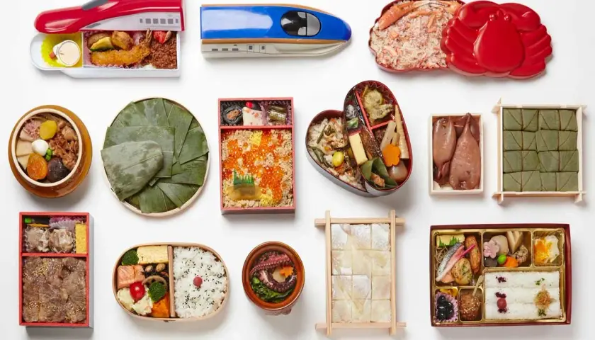 A variety of Japanese bento boxes are displayed, including train-shaped ones, a heart-shaped bento, and those filled with seafood, rice, and other delicacies.