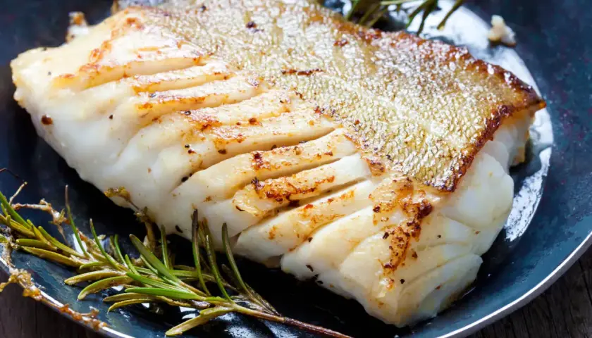 Un filet de poisson blanc parfaitement cuit, peau dorée et chair nacrée, servi avec du romarin frais. Une belle illustration de comment cuire du poisson blanc.