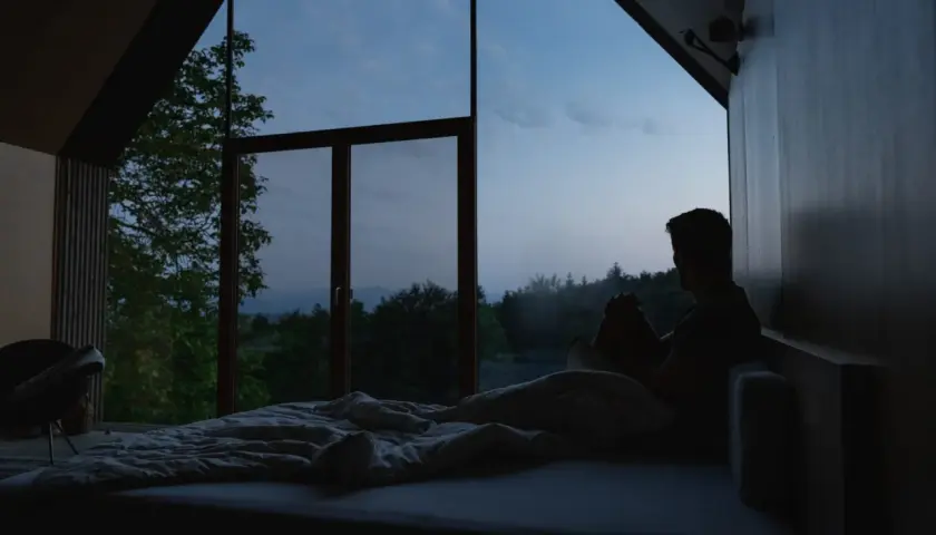 Mann sitzt im Bett, blickt aus dem Fenster in die Dämmerung. Ein ruhiger Moment, der den schlaf wach rhythmus widerspiegelt.