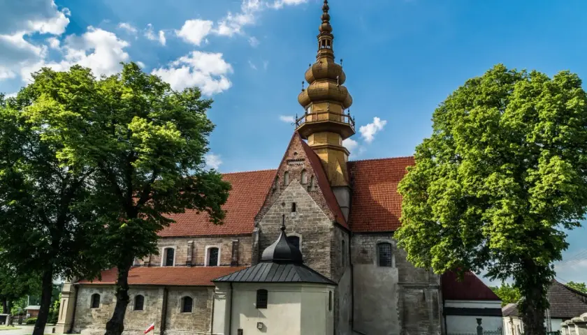 Kamienna świątynia z żółtą wieżą i czerwonym dachem, otoczona zielenią. W oddali widać fragmenty zabudowań, przypominające krajobraz wokół **zamku Krzyżtopór**.