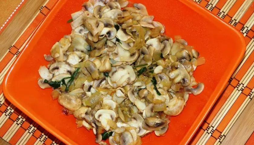 Champiñones salteados con cebolla y espinacas, servidos en un plato naranja sobre un mantel de bambú.