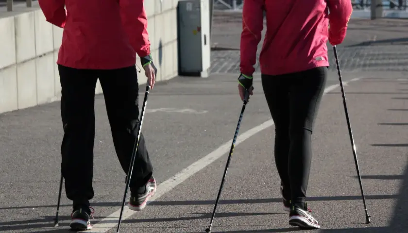 Dwie osoby w czerwonych kurtkach i czarnych spodniach idą z kijkami do nordic walking. Pokazują, jak ustawić kijki do chodzenia.