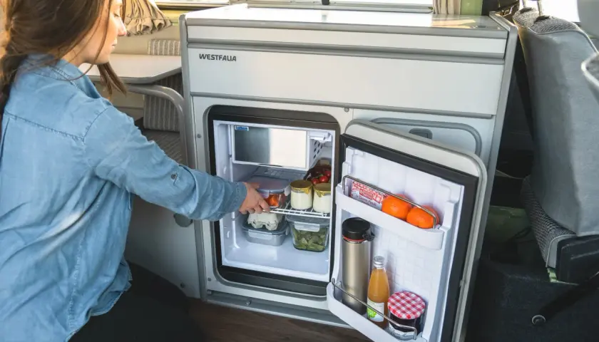 Une femme range des aliments dans un frigo pour van. Il contient des œufs, des légumes, des fruits et des boissons.