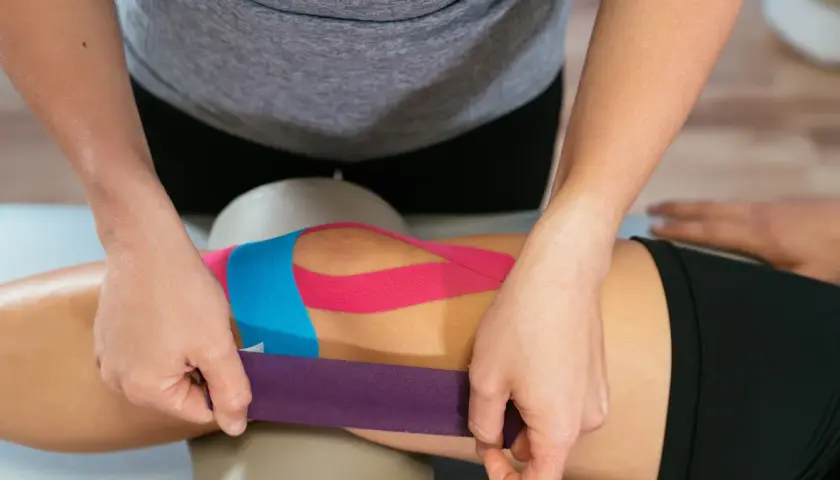 Fizjoterapeuta aplikuje kolorowe taśmy kinezyjne na kolano pacjenta, wspierając jego rehabilitację.