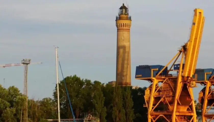 Najwyższa latarnia morska w Polsce, strzegąca portu, góruje nad żółtymi dźwigami i zielenią.
