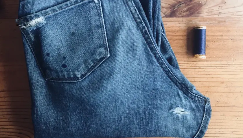A pair of blue jeans with a small rip on the back pocket and a spool of navy thread, ready for how to patch jeans.