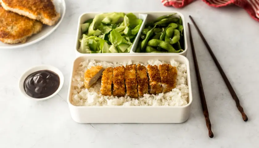 A delicious pork katsu bento box with rice, salad, edamame, and sauce.