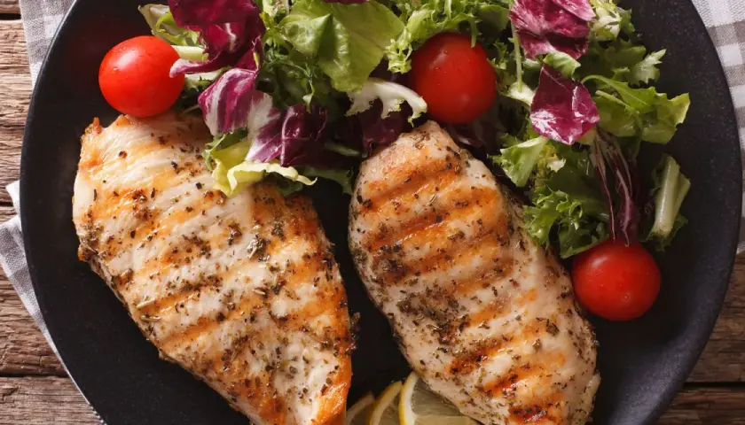 Due petti di pollo grigliati, conditi con erbe aromatiche, serviti con un'insalata fresca, pomodorini e spicchi di limone.