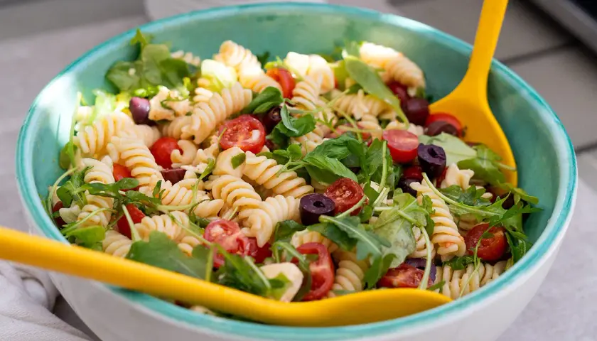Ensalada de pasta espectacular con fusilli, tomates cherry, aceitunas negras y rúcula, servida en un cuenco azul con cucharas amarillas.