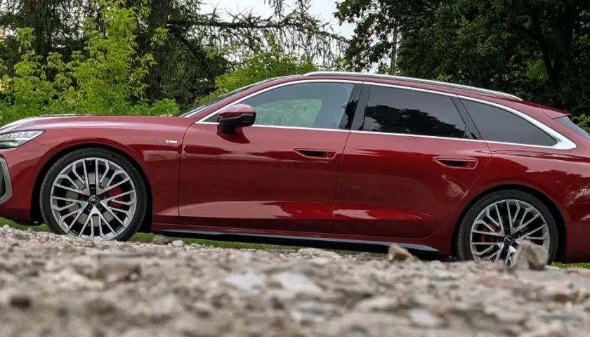 Lśniące, czerwone Audi A6 C9 Avant stoi na kamienistym podłożu, otoczone zielenią.