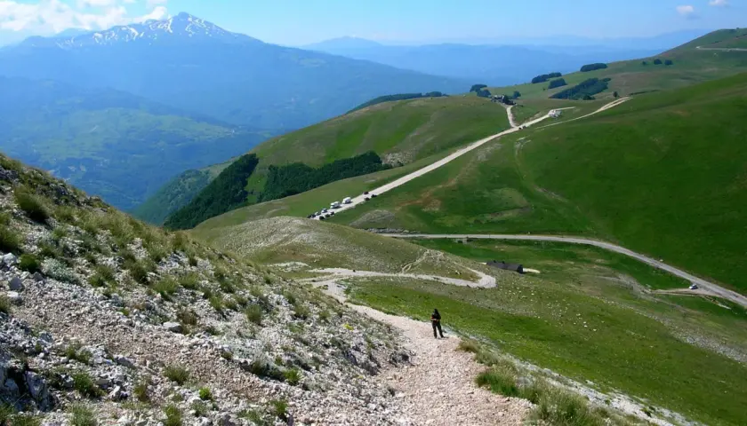 A person walks on a gravel path on a green mountain slope with a road winding through the hills and a snow-capped mountain in the distance.