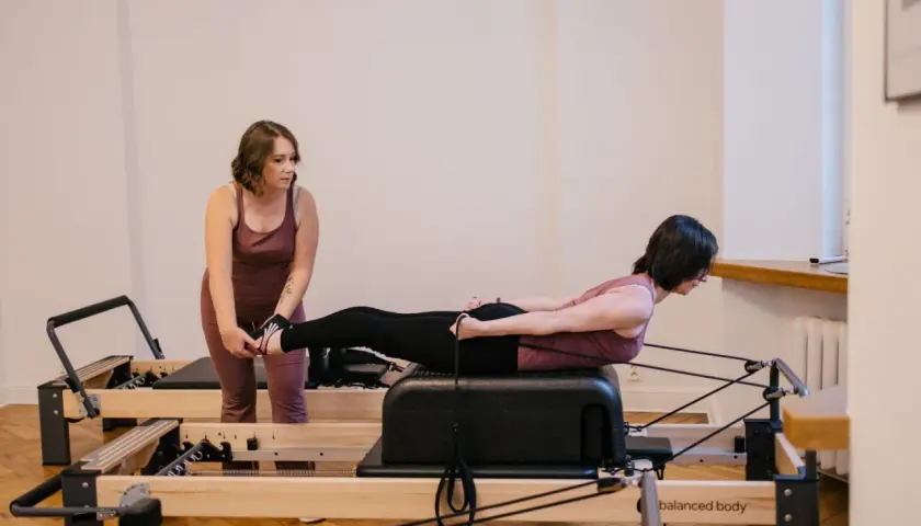 Instruktorka pomaga kobiecie w ćwiczeniu na reformerze. To pokazuje, na czym polega pilates: precyzja, kontrola i siła.
