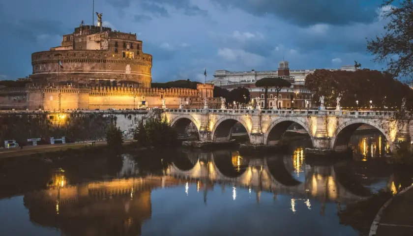 Le Castel Sant'Angelo, illuminé la nuit, se reflète dans le Tibre, avec un pont arqué en avant-plan.
