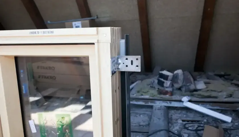 Drewniane okno dachowe z metalowym kątownikiem, gotowe do montażu. W tle widać materiały budowlane i elementy konstrukcji poddasza.