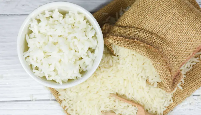 Ciotola di riso bianco cotto accanto a un sacco di riso crudo. Il riso crudo è noto per i suoi rimedi naturali.