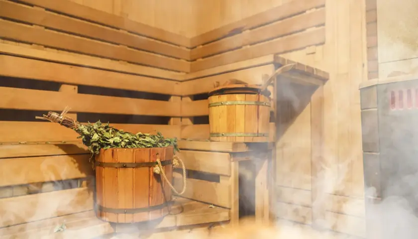 Drewniana sauna z miotełką i kadzią, idealna na zakwasy po treningu. Para unosi się, tworząc relaksującą atmosferę.