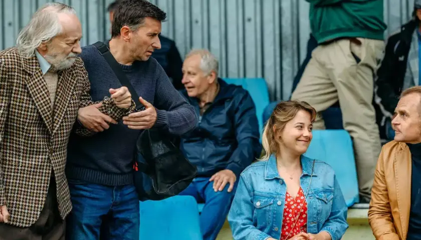 Grupa ludzi na trybunach, wśród nich starszy pan w tweedowej marynarce, wspierany przez młodszego mężczyznę. Scena jak z najlepszych polskich filmów.