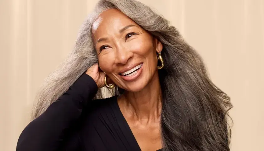 Mujer sonríe con cabello largo y ondulado, mostrando una hermosa transición a canas.