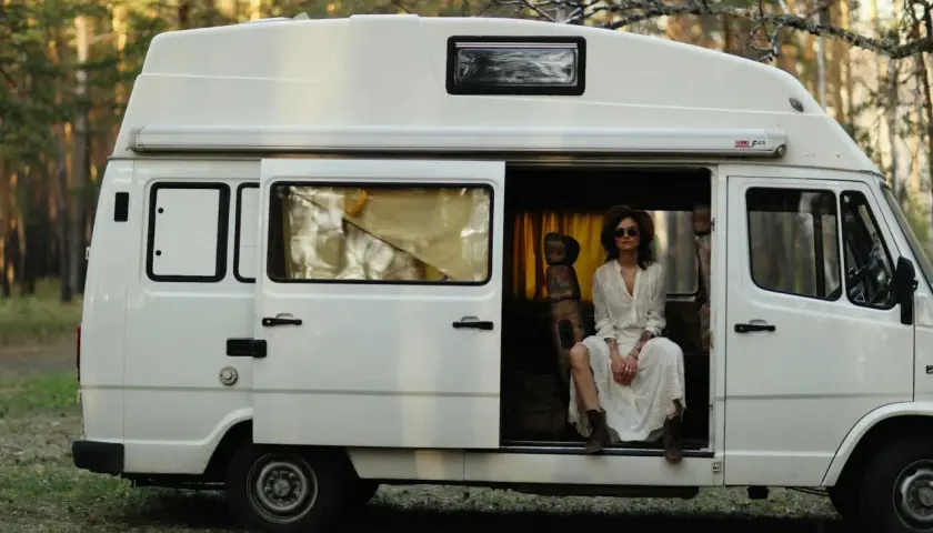 Kobieta w białej sukience siedzi w otwartych drzwiach białego kampera, otoczona lasem. Idealne polskie caravaning miejscówki na wakacje.