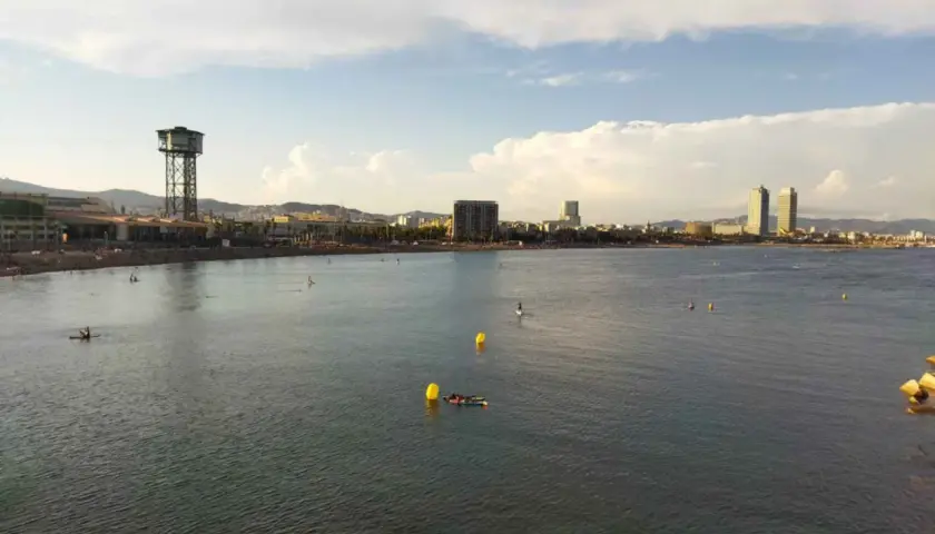 Vista de la playa y el mar con el icónico hotel vela al fondo. Personas disfrutan del agua en tablas de paddle surf.