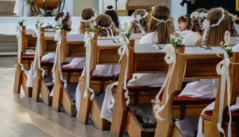 Dzieci w bieli, z wiankami na głowach, siedzą w ławkach kościelnych. Czekają na ważny moment, zastanawiając się, ile trwa komunia.