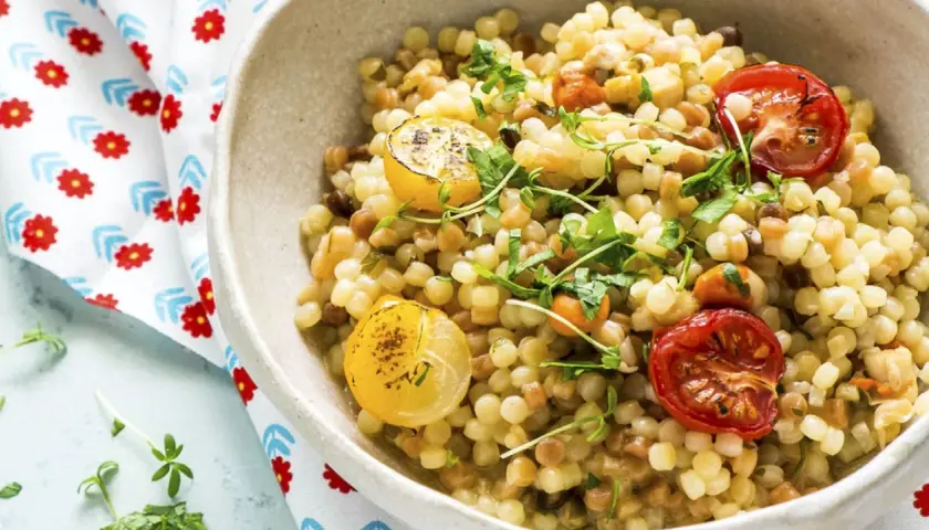 Un plato de pasta pequeña, que es semola, con tomates cherry asados y rodajas de limón, adornado con hierbas frescas.