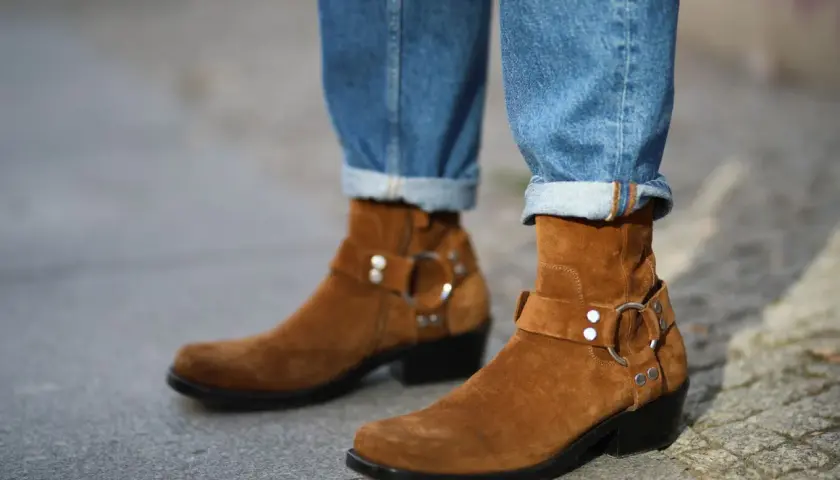 Botas de gamuza marrón con hebillas y jeans doblados. La piel girada de las botas contrasta con el denim.
