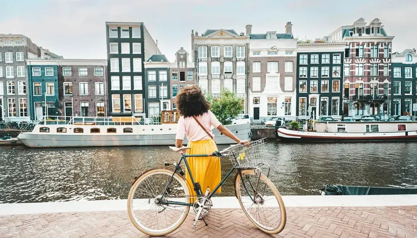 Kobieta z rowerem na tle amsterdamskich kamienic i kanału. Idealne miejsce na odkrywanie tras rowerowych Europa.