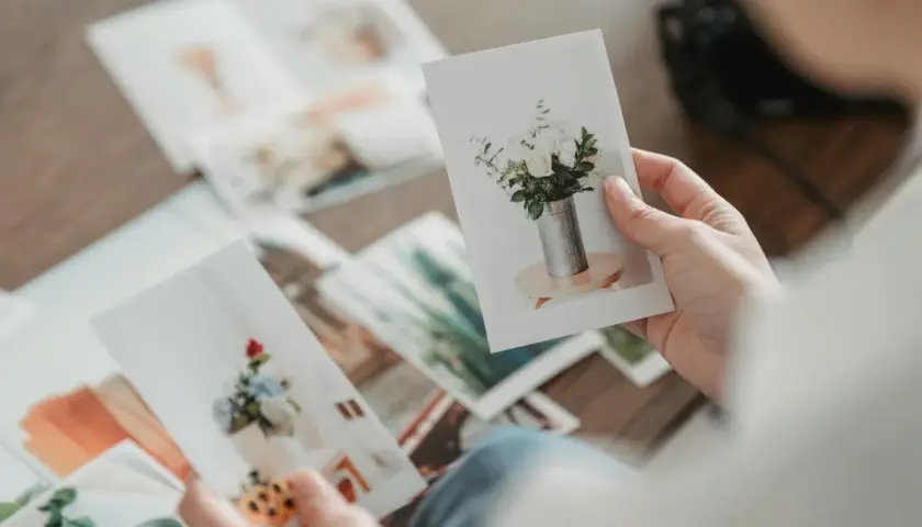 Dłonie przeglądają zdjęcia na papierze kredowym, ukazujące bukiety kwiatów w wazonach. To wspomnienia utrwalone na papierze fotograficznym.