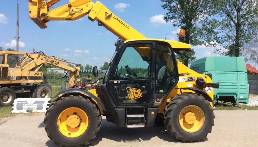 Żółty ładowarka teleskopowa JCB 531-70 Loadall stoi na placu. W tle koparka CAT i zielona kabina ciężarówki.
