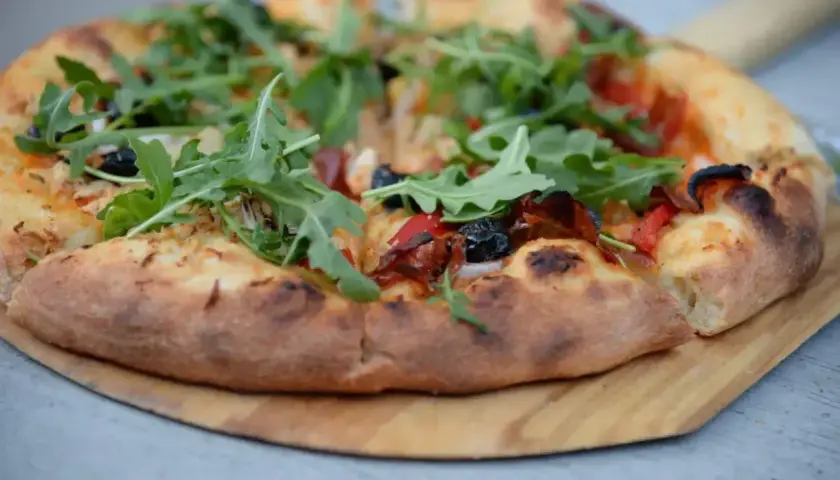 Pyszne ciasto na pizzę z rukolą, oliwkami i papryką, podane na drewnianej desce.