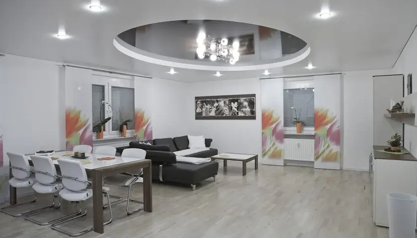 Nowoczesny salon z okrągłym sufitem napinanym, który odbija elegancki żyrandol. Wygodna kanapa i stół z krzesłami tworzą przytulną przestrzeń.