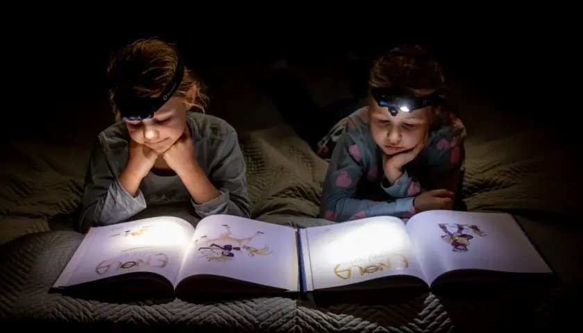 Dwie dziewczynki z latarkami na głowach czytają książkę w ciemności. To świetny sposób, by przygotować się na blackout i nie nudzić się!