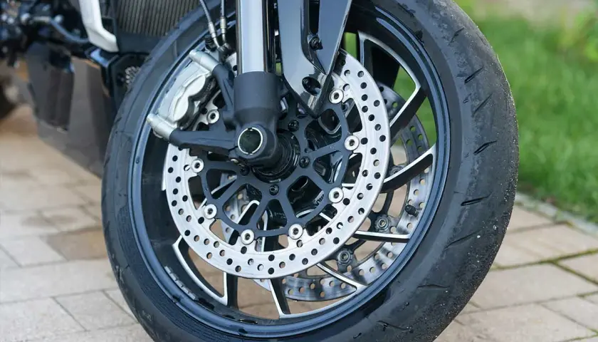 Primer plano de la rueda delantera de una motocicleta, con discos de freno perforados y pinzas. Se ve el neumático desgastado, listo para reparar rueda.