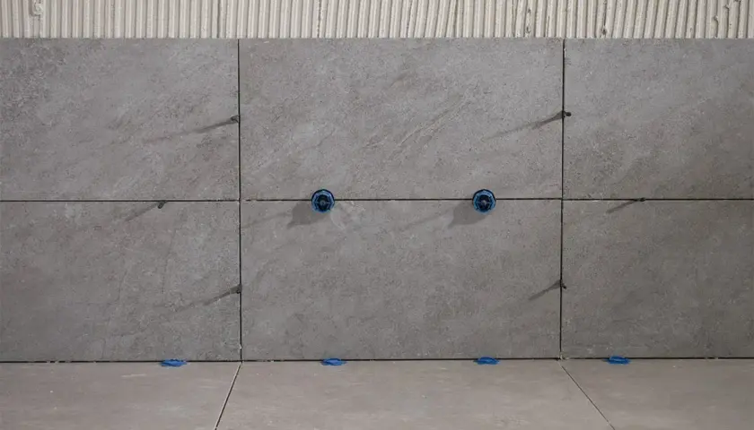 Posa di piastrelle grigie con distanziatori blu. Le fughe piastrelle non rettificate sono visibili, con adesivo sottostante.