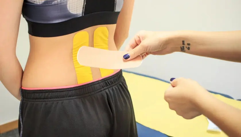 Naklejanie tejpy na plecy w odcinku lędźwiowym. Dłoń z tatuażem "You can do it" przygotowuje plaster.