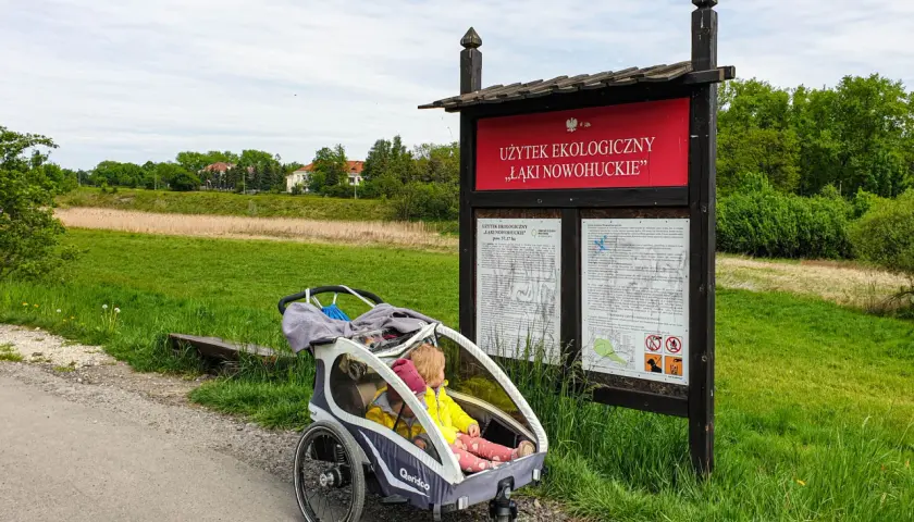 Dzieci w wózku czekają na spacer po malowniczych łąkach nowohuckich, obok tablicy informacyjnej.