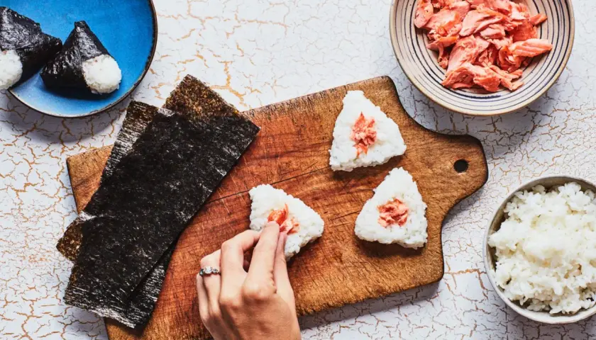 Making onigiri rice recipe: hands shaping rice balls with salmon filling, nori sheets, and bowls of rice and salmon.