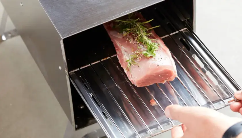 Un filet de porc fumé, assaisonné d'herbes et de poivre, est placé sur une grille dans un fumoir en acier inoxydable.