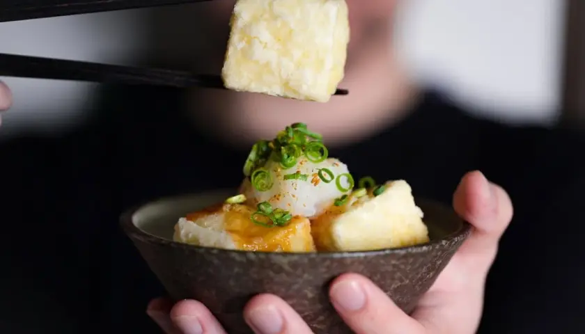 Chopsticks lift a piece of crispy age tofu, ready to be dipped into a savory sauce.