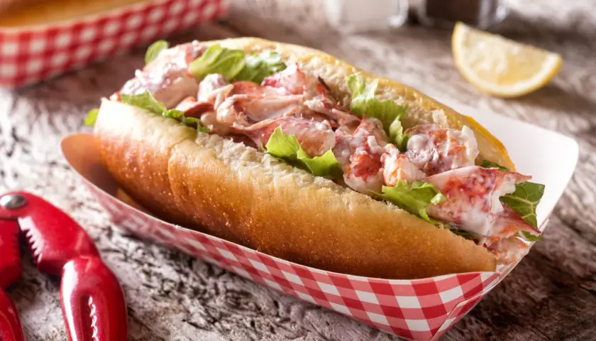 Pyszny lobster roll z kawałkami homara i sałatą w bułce, podany w czerwono-białym papierowym opakowaniu. Obok szczypce do homara.