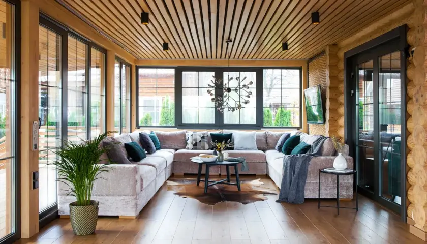 Aménagement véranda moderne avec grand canapé modulable, table basse, tapis peau de vache et plantes vertes. Lumière naturelle abondante.