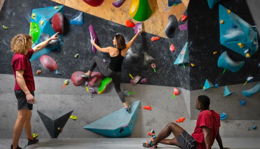 Drei Personen bouldern an einer bunten Wand. Ein Mann gibt Tipps, eine Frau klettert, ein Mann ruht.