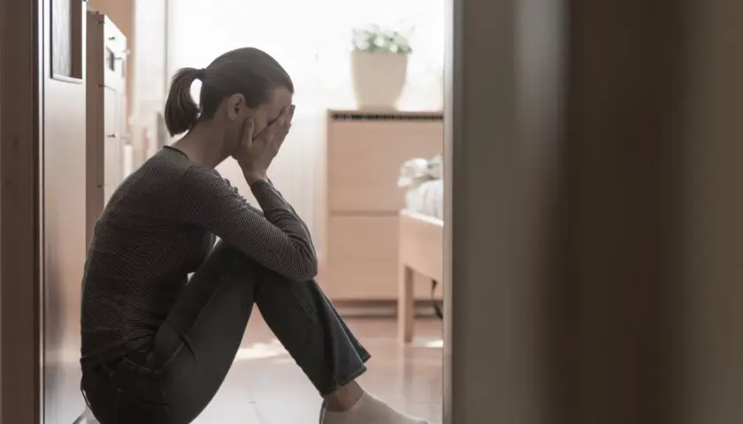 Kobieta siedzi na podłodze, zakrywając twarz dłońmi. W tle widać łóżko i roślinę. To obraz smutku, być może baby blues.