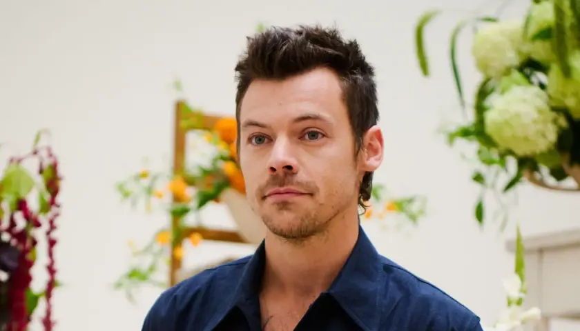 Harry Styles con un moderno mini mullet, mirada intensa y camisa azul, rodeado de arreglos florales.