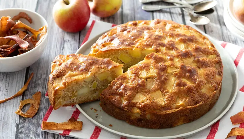 Un délicieux gâteau aux pommes, doré et moelleux, avec des morceaux de fruits visibles. Des pommes fraîches et des chips de pommes complètent la scène.