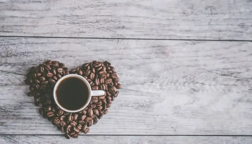 Una tazzina di caffè nero, circondata da chicchi di caffè disposti a forma di cuore su un tavolo di legno. Un invito al caffè in allattamento.