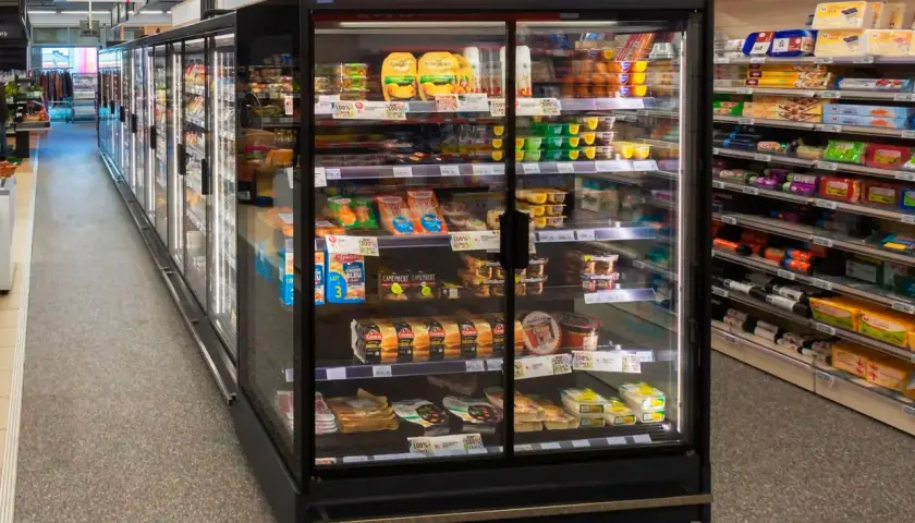 Chłodnia z produktami spożywczymi w supermarkecie, z widocznymi serami, wędlinami i deserami.